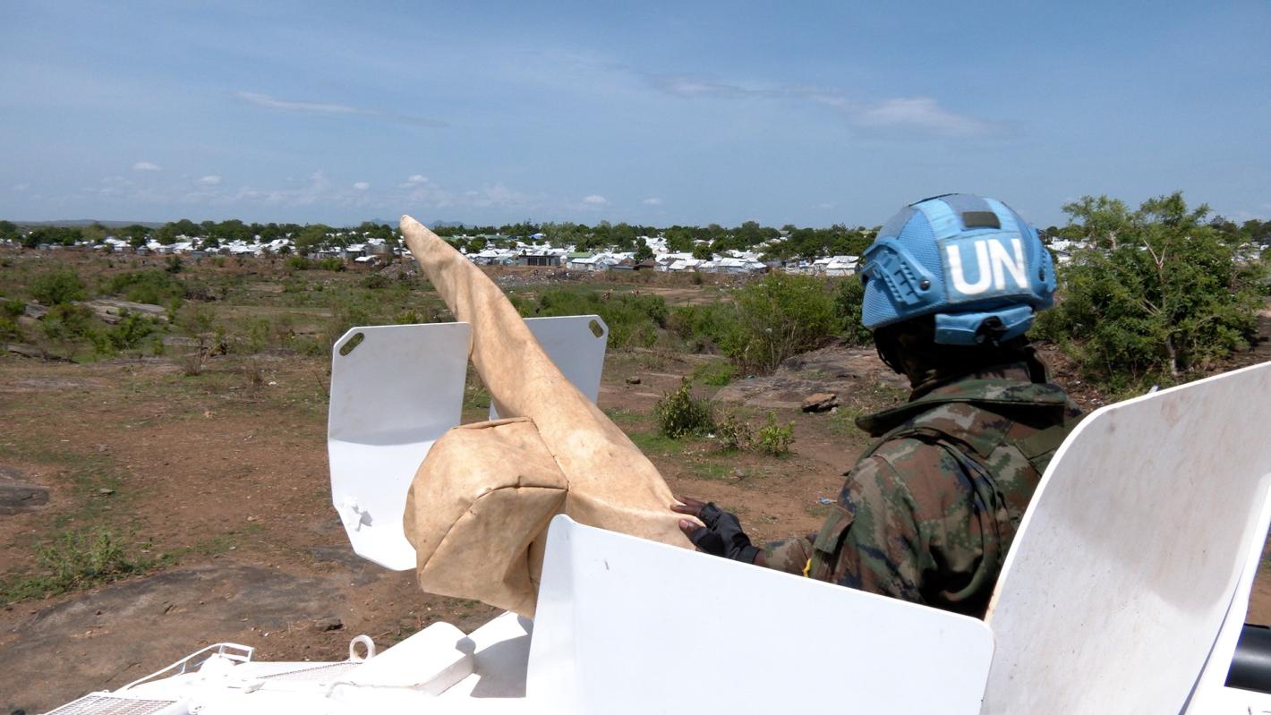 Rwandan peacekeepers patrol around camps for displaced communities to strengthen security as political tensions rise and clashes continue between armed forces in pockets of the country. A peacekeeper peering out of the top of a tank across a grassy field. The peacekeeper is facing away from the camera.