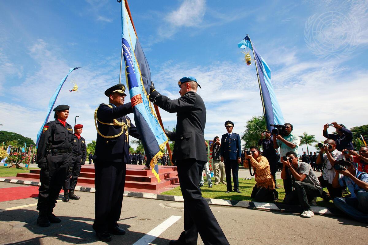 A UN peacekeeper hands over a ceremonial flag to a Timor-Leste police officer during a formal event, as photographers and onlookers capture the moment under a clear blue sky.