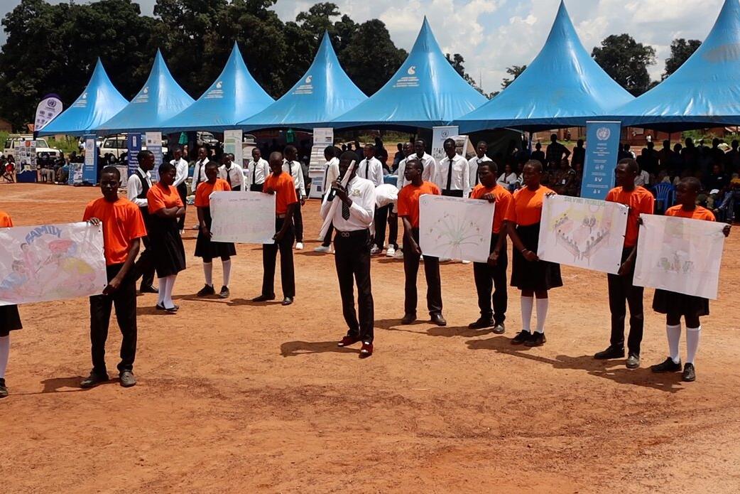 Students in orange shirts and black skirts or trousers hold large hand-drawn posters during an outdoor event with blue tents and a crowd in the background.