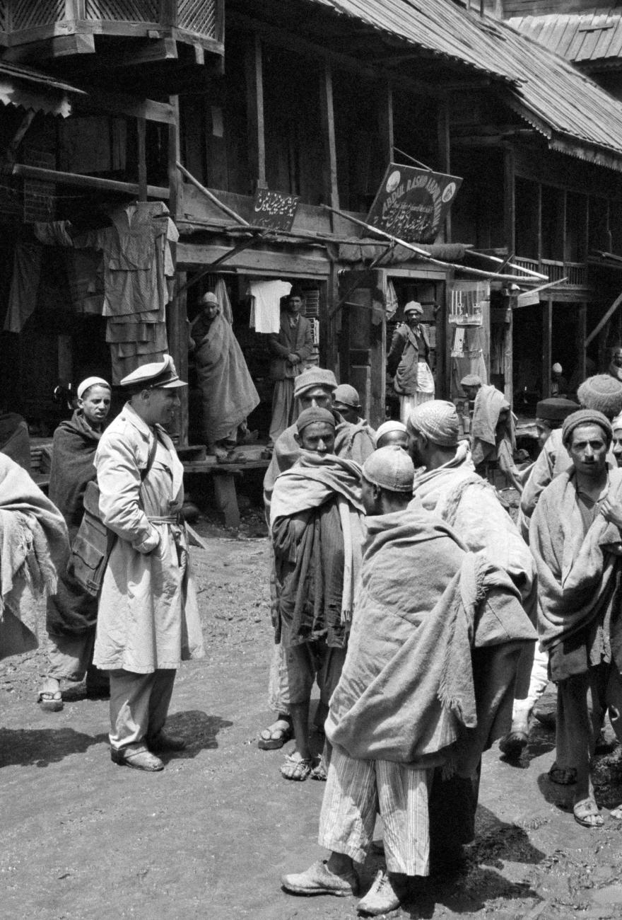 UN military observer in uniform speaks with a group of people wrapped in blankets in a rural outdoor setting with wooden structures in the background.