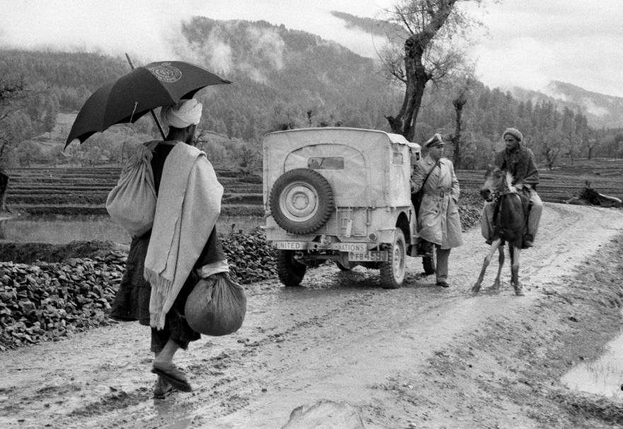 UN jeep on a muddy road with two people nearby; one rides a horse while another walks carrying an umbrella and bags.